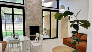 A modern living room with a stone fireplace, a dining area with white furniture, and a large window overlooking a garden. - Brand New 2x2 Escape | Relax & Unwind (Herron)