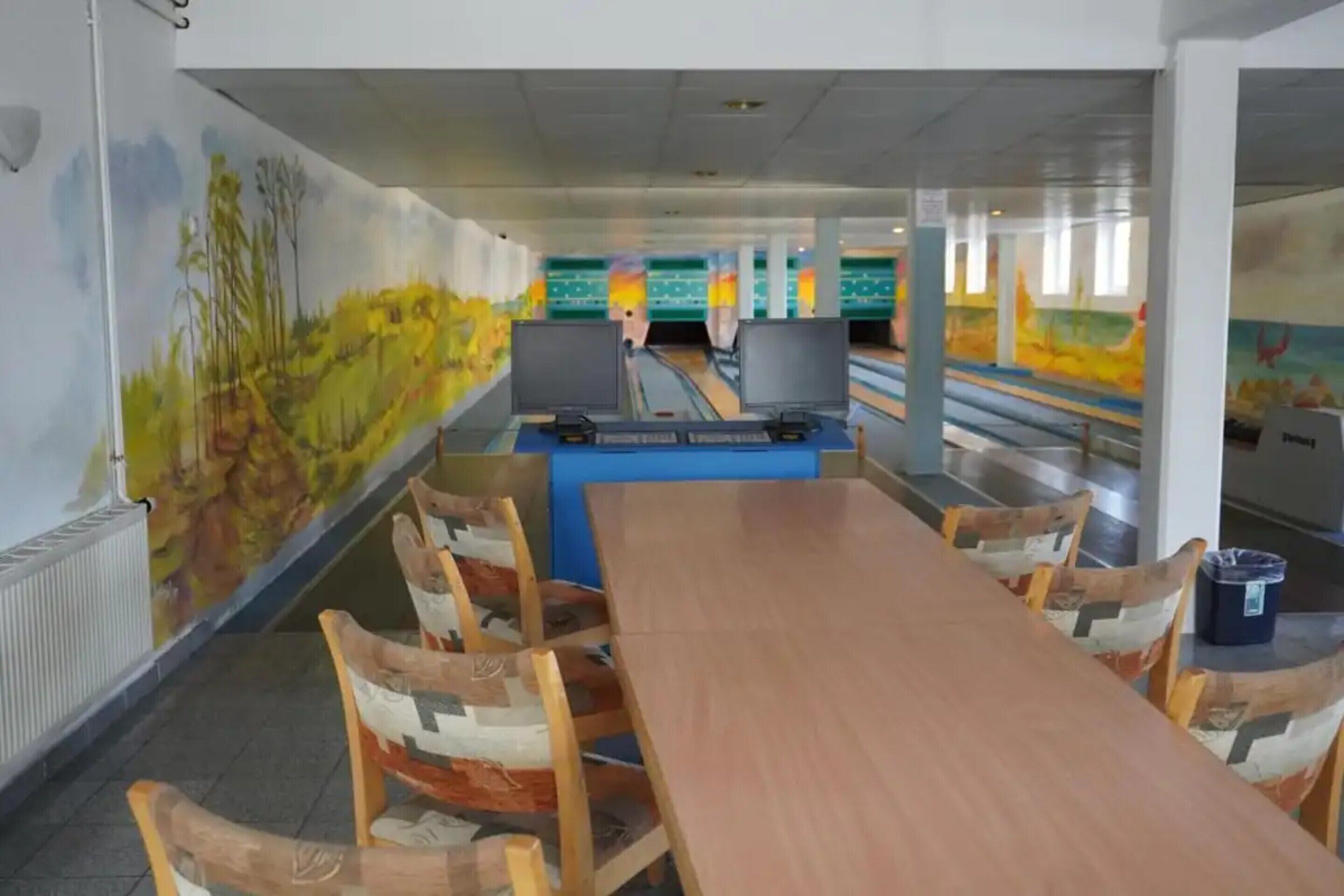Una bolera con mesas y sillas de madera, una pista de boliche y un mural en la pared.