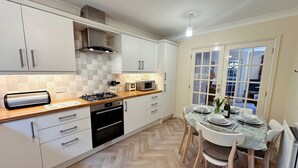 A modern kitchen with white cabinets, a herringbone floor, a gas stove, and a dining area with a table set for two. - Tranquil retreat in idyllic Wooler, Northumberland National Park, pet friendly (Wooler)