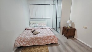 A bedroom with a bed, a nightstand, a lamp, a window with blinds, and a mirror. - Spacious 3‑Bedroom | Family & Work Stay | Near Campbelltown Hospital (Airds)