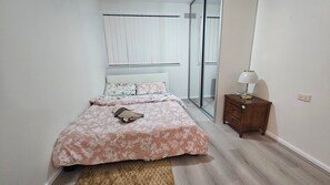 A bedroom with a bed, a nightstand, a lamp, a window with blinds, and a mirror. - Spacious 3BR Home for Family & Work Stays Near Campbelltown | Parking Fast WiFi (Airds)