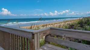 Terraza de madera con vistas a una playa, olas, arena y vegetación escasa.
