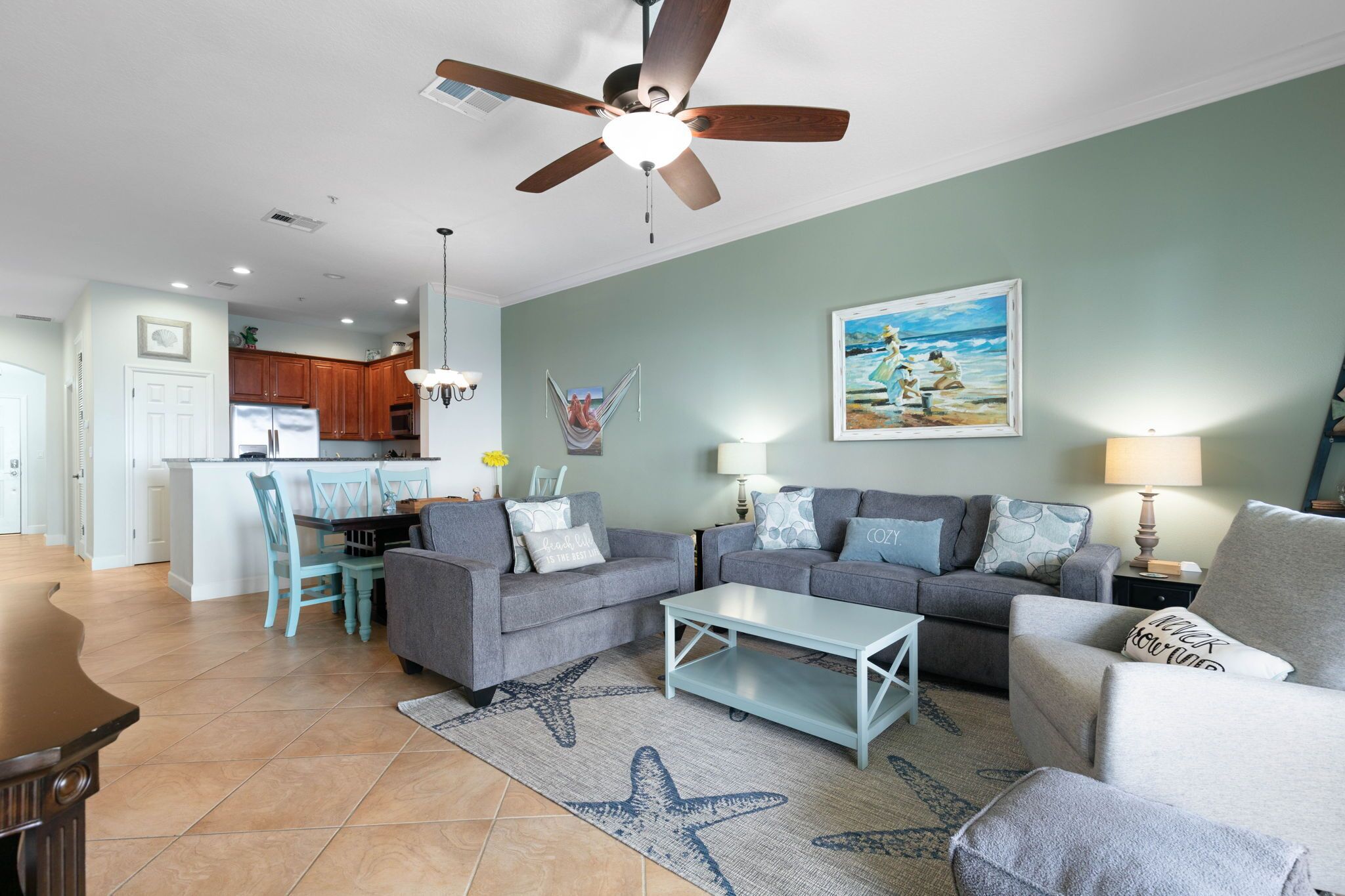 A living room with a ceiling fan, a dining area with a table and chairs, and a kitchen in the background.