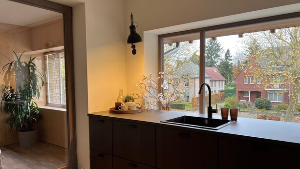 A modern kitchen with a large window, a black pendant light, and a view of a red brick house. - Stylish Apartment in Bree Centre (Bree)