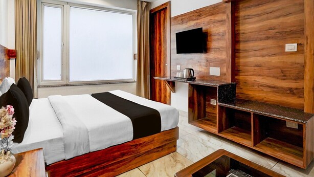 Quarto de hotel com cama, televisão fixada numa parede de madeira, cafeteira e janela com cortinas.