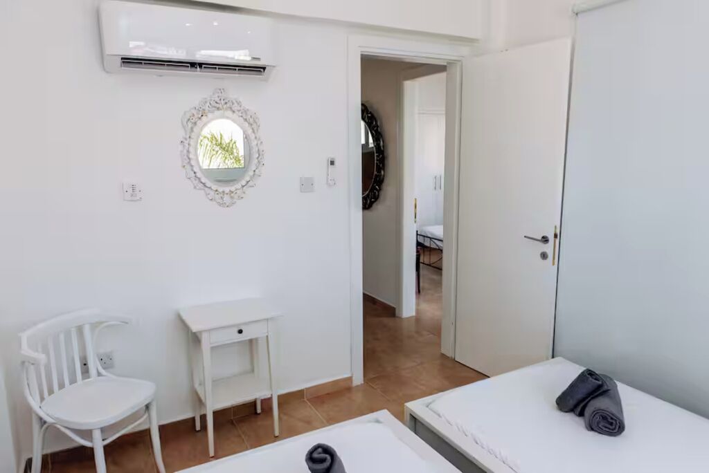 Un dormitorio con cama, una silla, una mesita, un espejo y un aire acondicionado.