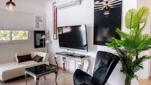 Ein modernes Wohnzimmer mit einem weißen Sofa, einem schwarzen Ledersessel, einem Glastisch, einem Flachbildfernseher und einer Topfpflanze.