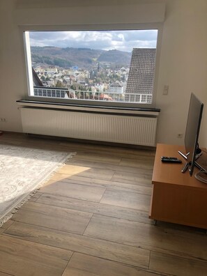 Ein Zimmer mit großem Fenster, Heizkörper, Holzboden und Schreibtisch mit Computer und Tastatur.