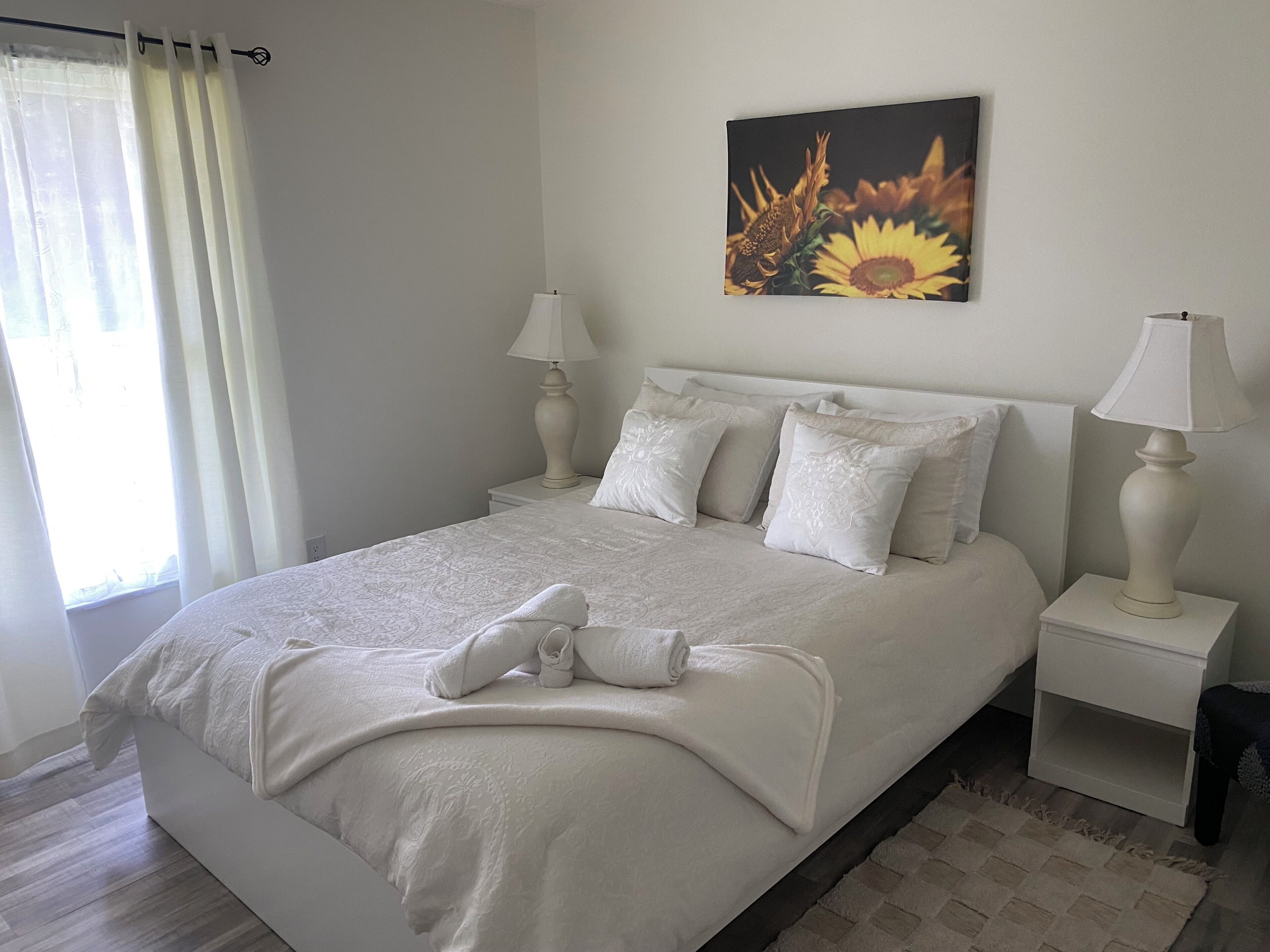 Una cama bien hecha con edredón y almohadas blancos, un cuadro de girasoles en la pared y una ventana con cortinas.