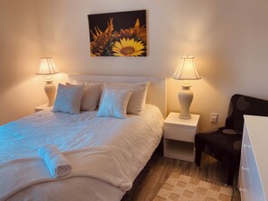 A neatly made bed with a rolled towel, two bedside lamps, a wall-mounted painting of sunflowers, and a chair with a patterned cushion. - Cozy 1-bedroom apartment in peaceful Port Charlotte with AC. Close to amenities. (Port Charlotte)