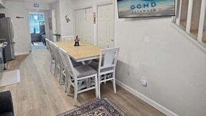 Un comedor con una mesa de madera y sillas blancas, una cocina con refrigerador y un televisor de pared mostrando 'OCMD GETAWAYS'.