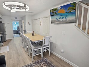 Un coin repas avec une table en bois et des chaises blanches, une cuisine équipée d’un réfrigérateur, et un téléviseur fixé au mur qui diffuse l’émission « OCMD GETAWAYS ».