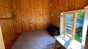 Um pequeno quarto de madeira com uma cama, uma janela com moldura de metal e uma estante com um livro.