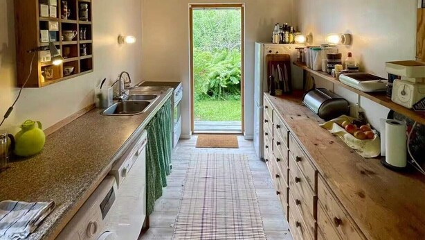 Uma cozinha compacta com vigas de madeira, máquina de lavar roupa e vista através de uma porta para um jardim.