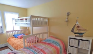 Un lit superposé pour enfant avec une couvre-lit colorée, une table de chevet blanche avec des paniers, et une décoration murale représentant un voilier.