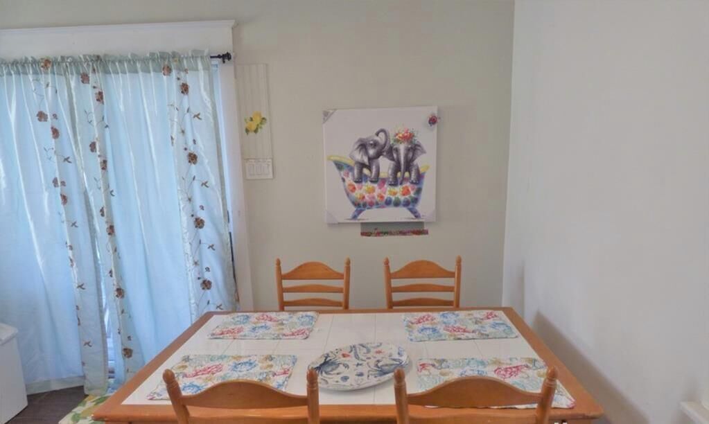 Een eethoek met een tafel voor vier personen, houten stoelen, een tafelkleed met bloemenprint en een aan de muur hangend schilderij van olifanten.
