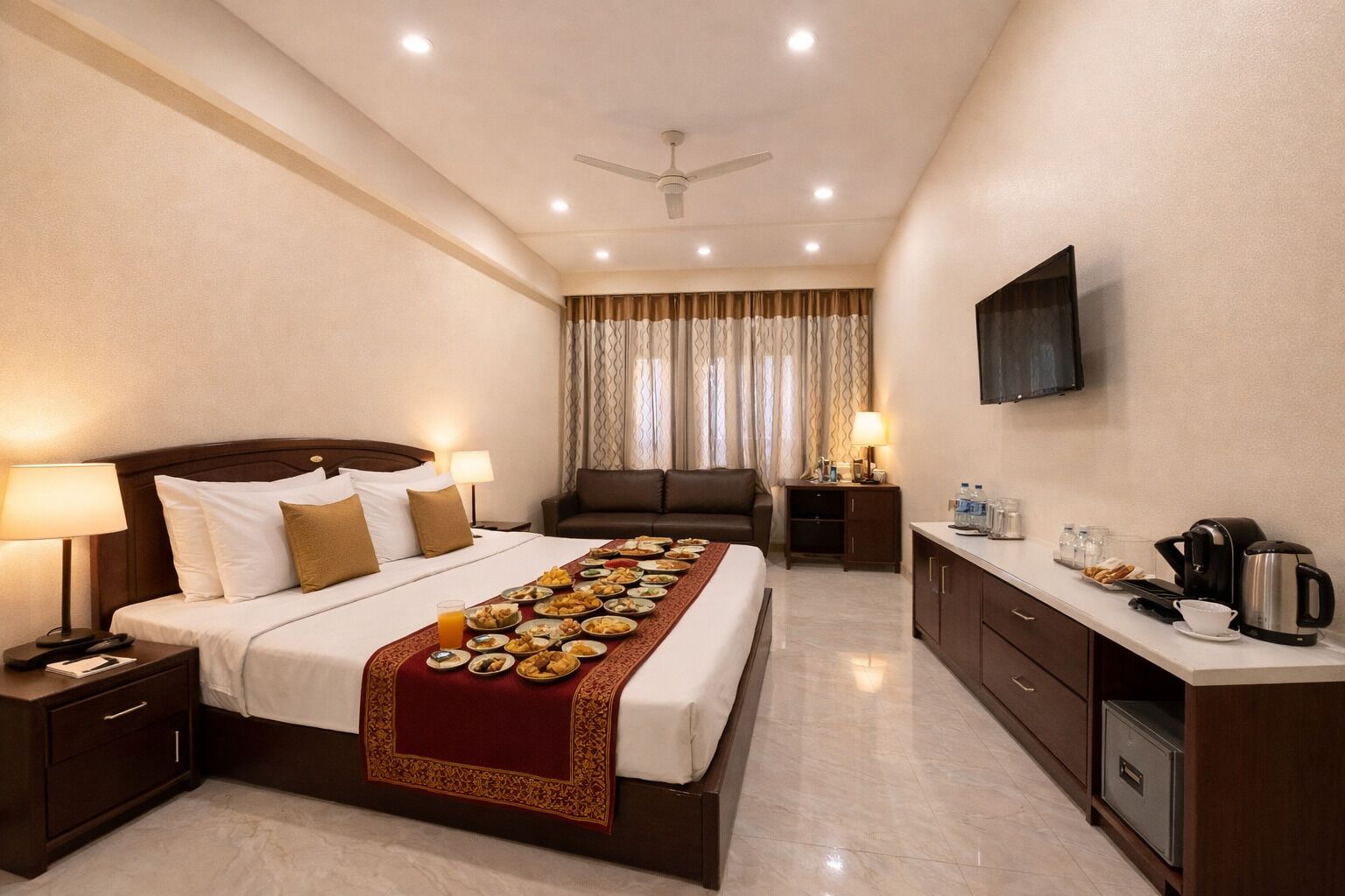 Habitación de hotel con cama, mesitas de noche, sofá, televisor, escritorio con cafetera y zona de cocina.