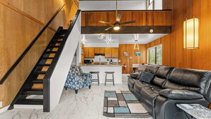 Un salon moderne avec un canapé en cuir noir, un tapis à motifs, un escalier et, au fond, un coin cuisine.