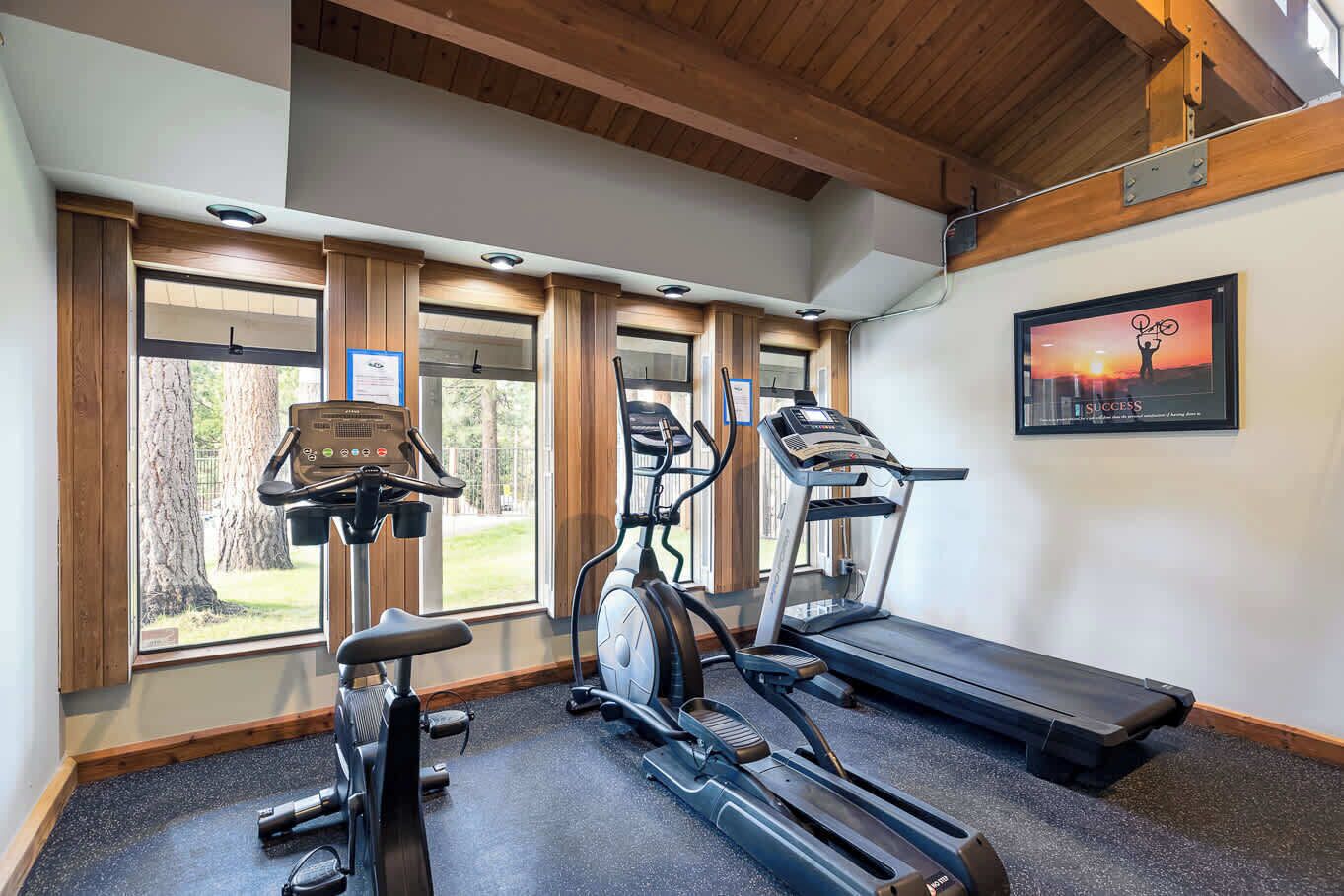 A gym with cardio machines, a treadmill, and a large window with a view of trees.