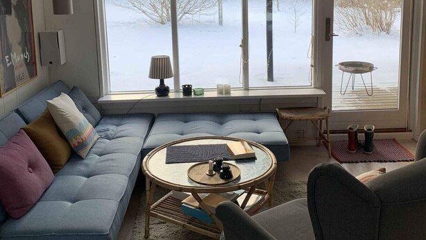 A living room with a sectional sofa, a round coffee table, and a chair. There are large windows with a view of a snowy landscape.