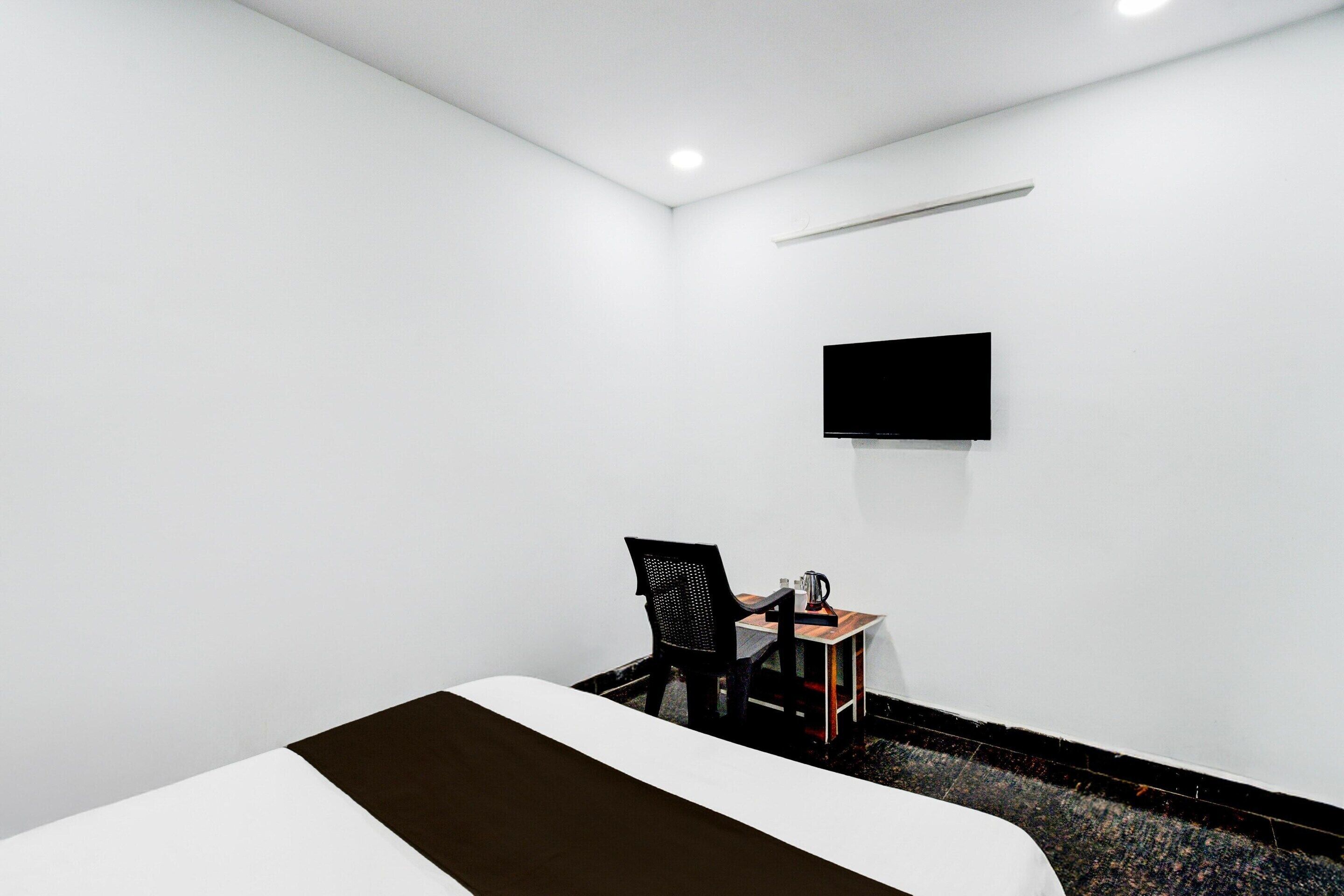 A hotel room with a bed, a chair, a small table, a flat-screen TV mounted on the wall, and a ceiling light.