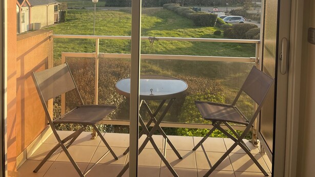 A balcony with two chairs and a small table, offering a view of a grassy field and distant buildings. - Sea view apartment facing the beach (Clohars-Carnoët)