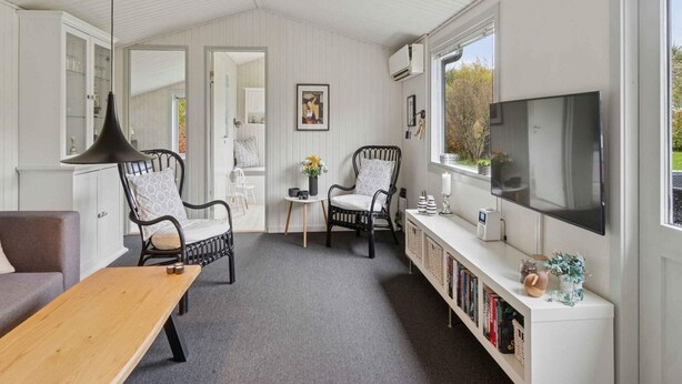 Ein gemütliches Zimmer mit einem Holztisch, zwei schwarzen Stühlen, einem Bücherregal, einem Fernseher und einem Fenster mit Blick auf Bäume.