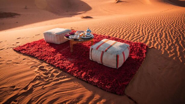Un montaje para picnic en el desierto con una mesa, cojines y una manta sobre la arena.