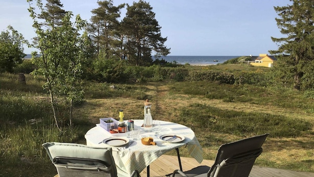 An outdoor dining area with a table set for two, surrounded by chairs, overlooking a beach and a yellow building. - 4 Star Holiday Home in Glesborg (Glesborg)