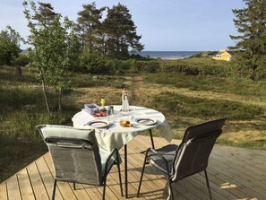 An outdoor dining area with a table set for two, surrounded by chairs, overlooking a beach and a yellow building. - 4 Star Holiday Home in Glesborg (Glesborg)