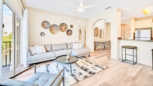 A spacious living area with a sofa, coffee table, and a view of the kitchen. - Upstay - Condo Near Disney w Pool-spa-sauna-games (Winter Garden)