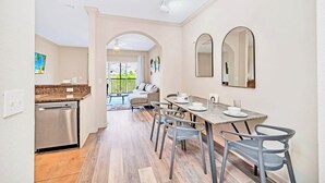 A modern kitchen with a dishwasher, a dining area with a table and chairs, and a view of the outside. - Upstay - Condo Near Disney w Pool-spa-sauna-games (Winter Garden)