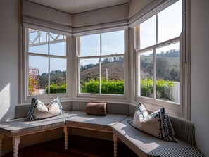 A bay window with three panes, a striped bench with cushions, and a view of a green landscape. - Stunning Dartmouth Views | Stylish Coastal Home (Dartmouth)