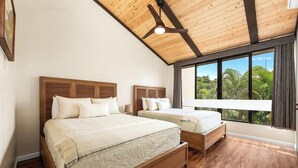 Ein Schlafzimmer mit zwei Betten, Holz-Kopfteilen, einem Deckenventilator und großen Fenstern mit Blick auf Grünflächen.