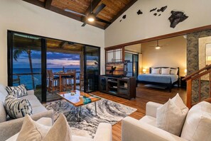 A spacious living area with a large window overlooking the ocean, a dining area, a bedroom with a bed and nightstand, and a staircase leading to another level. - Kksr#4303 Direct Oceanfront, Top Floor, Ac, Remodeled, Sleeps 8 (KAILUA KONA)