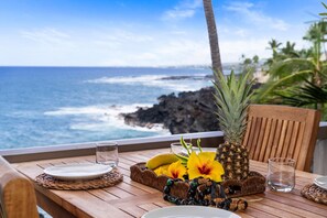 A wooden table with plates, glasses, a pineapple, bananas, and flowers by a window overlooking the ocean. - Kksr#4303 Direct Oceanfront, Top Floor, Ac, Remodeled, Sleeps 8 (KAILUA KONA)
