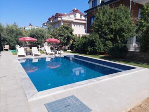 A rectangular swimming pool with a tiled edge, surrounded by a paved deck. There are lounge chairs and umbrellas, and a building with a red roof in the background. - Fethiyeyazevim (Fethiye)