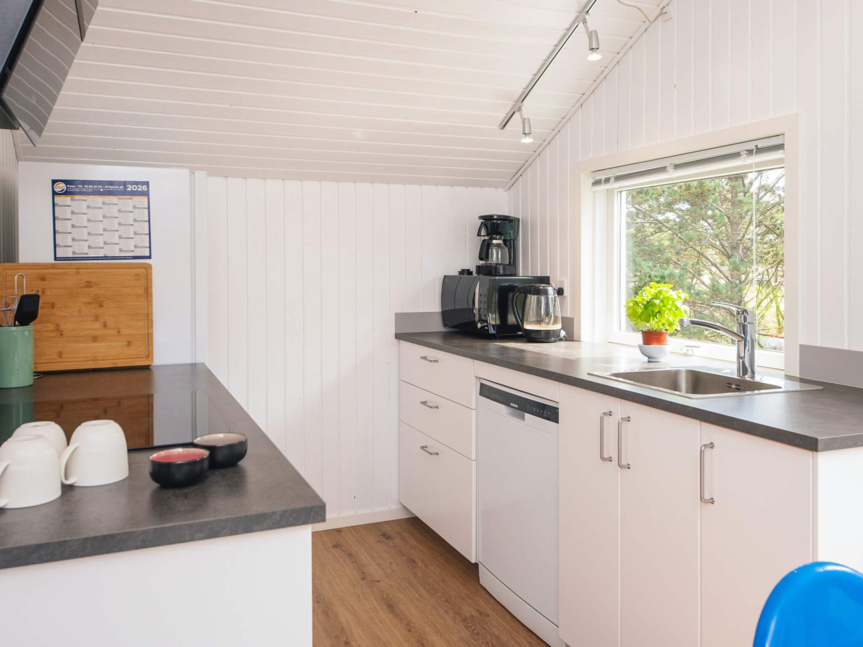 A modern kitchen with white cabinetry, a coffee maker, a sink, and a window with a view of trees.