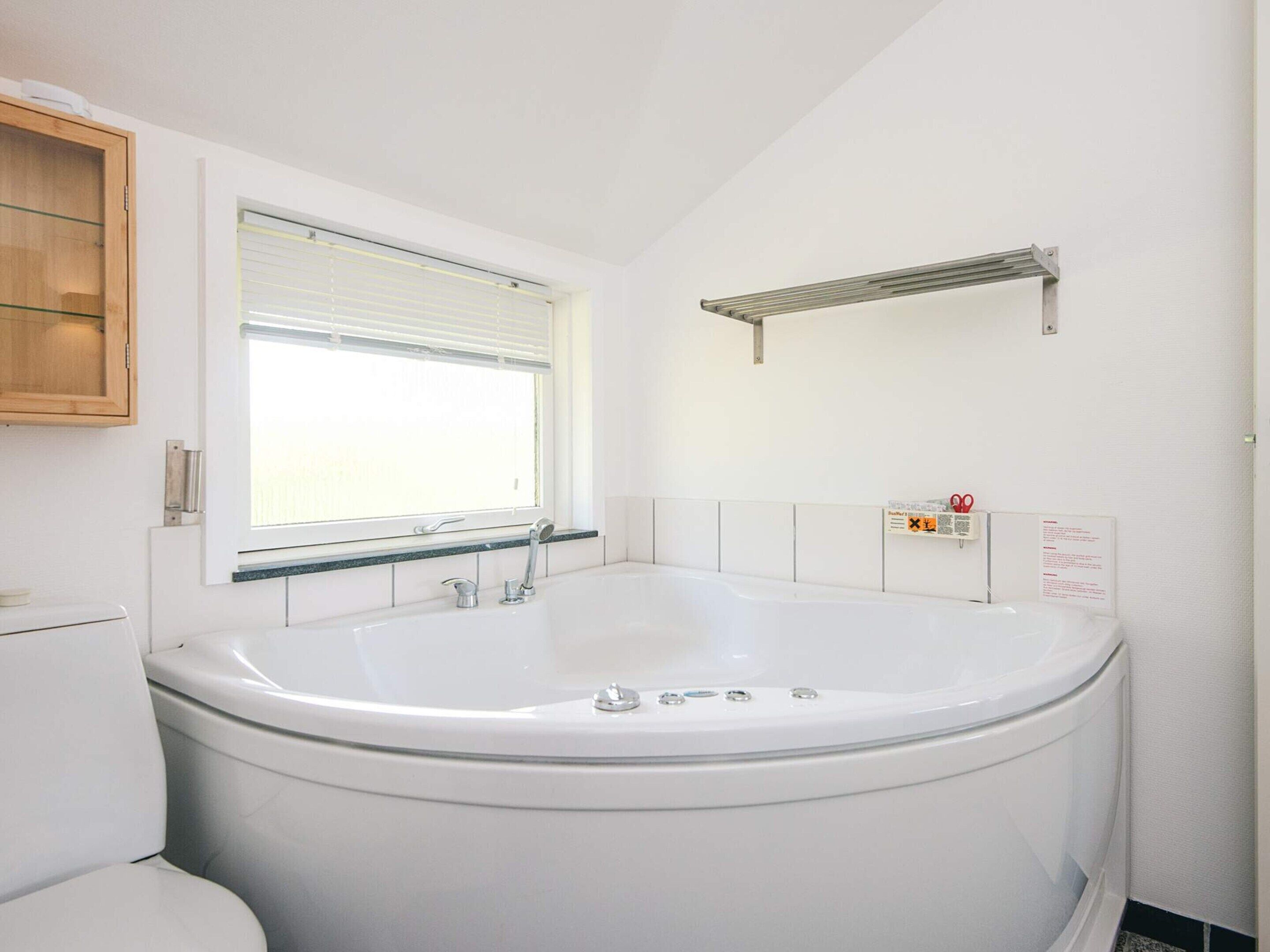 Une salle de bain avec une baignoire blanche, une fenêtre avec des stores, un meuble en bois et un porte-serviettes.