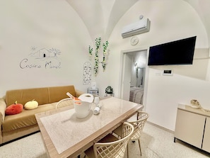 A modern living room with a sofa, a dining table, chairs, a TV, and an air conditioning unit. - Casina Mameli: cozy little house in the heart of Salento (Nardò)