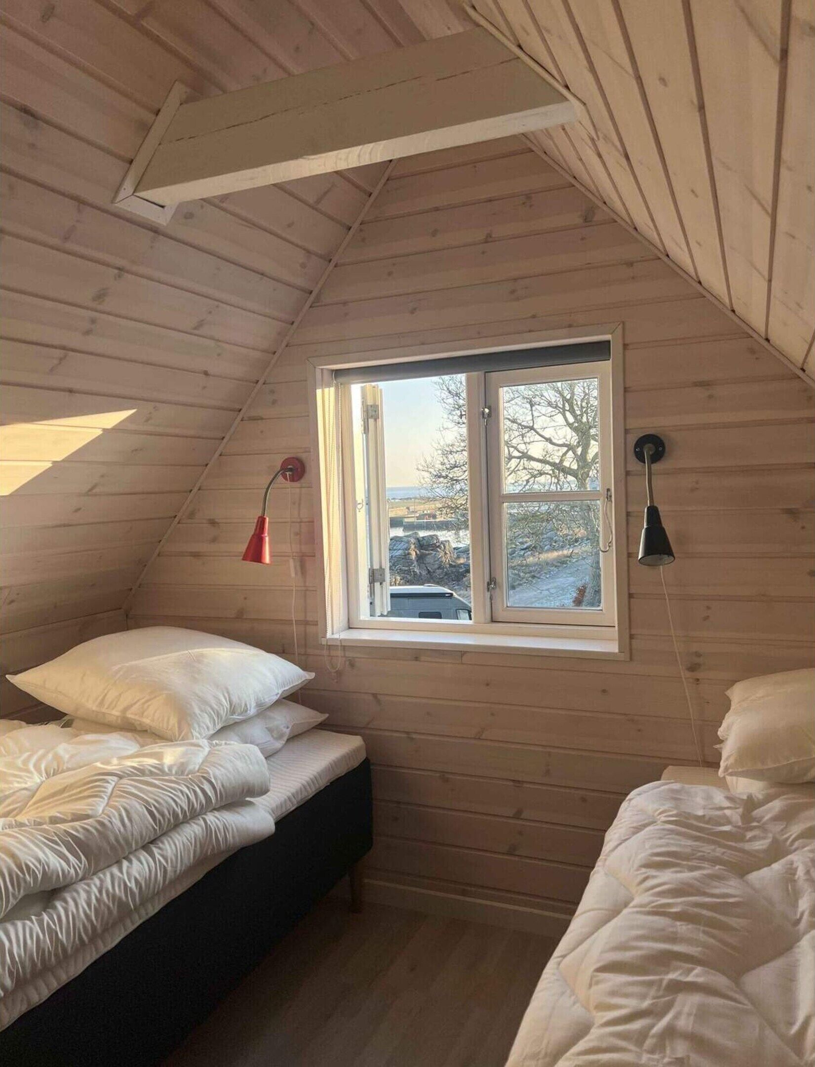 A wooden cabin room with a sloped ceiling, a window with a view, a bed with white bedding, and a red lamp.