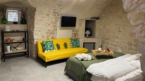 A room with a stone wall, a yellow sofa, a bed, a TV, and a shelf with items.