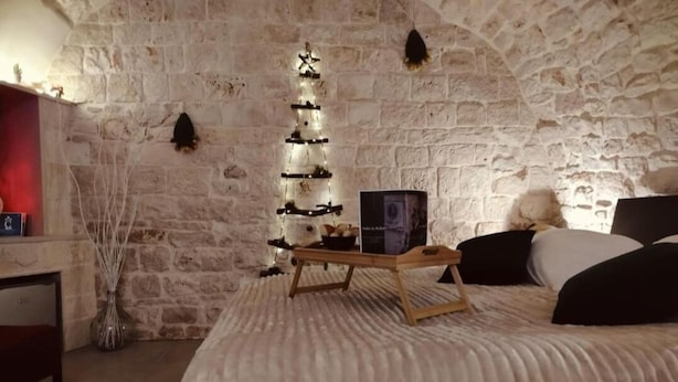 A stone-walled room with a bed, a wooden side table, a wall-mounted light fixture, and a small decorative item on the wall.