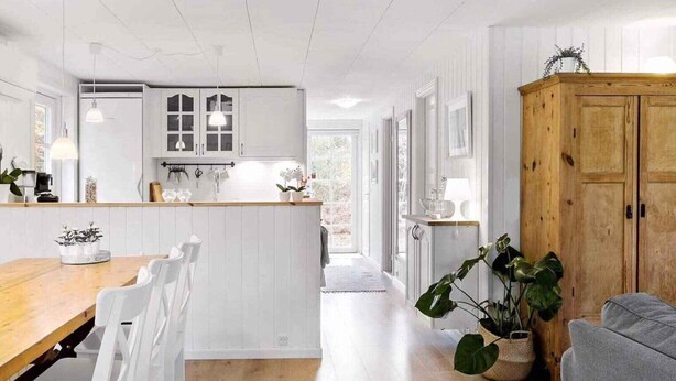 A modern kitchen with white cabinetry, a wooden dining table, and chairs. There is a large wooden cabinet and a potted plant in the living area. - 4 Star Holiday Home in Hadsund (Hadsund)