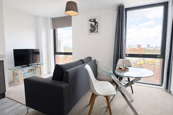 Ein modernes Wohnzimmer mit Sofa, Fernseher, Glastisch und großen Fenstern.