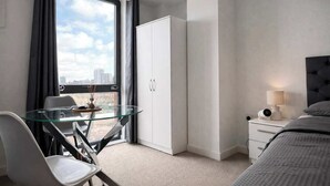 Moderni hotellihuone, jossa on lasinen ruokapöytä, valkoinen vaatekaappi, harmaalla päätyosalla varustettu sänky ja suuret ikkunat, joista avautuu näkymä kaupunkiin.