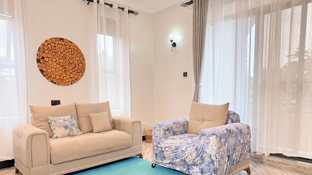 A living room with a beige sofa, a floral armchair, a blue rug, and a wall-mounted circular wooden decoration. - Constatia Homes (Kampala)