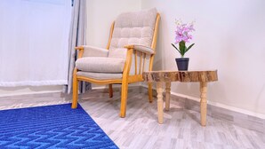 A room with a wooden chair, a small wooden table with a potted plant, a blue rug, and a window with curtains. - Constatia Homes (Kampala)