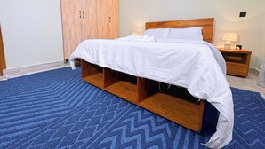 A bedroom with a bed, wooden wardrobe, nightstand, and a blue patterned carpet. - Constatia Homes (Kampala)