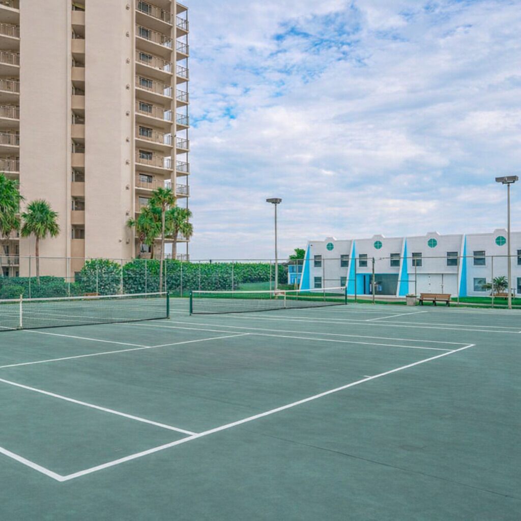 Una cancha de tenis con una red, rodeada por una valla, y contigua a un edificio de varios pisos con balcones.